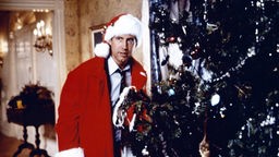 Chevy Chase steht im Film "Schöne Bescherung" als Weihnachtsmann verkleidet neben einem Weihnachtsbaum. 
