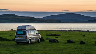 Im Abendlicht steht ein Campervan auf einer grünen Wiese zwischen Schafen direkt an einem idyllischen Platz am Meer. Grossbritannien.