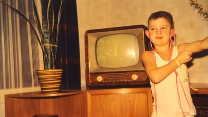 Ein Junge steht im Wohnzimmer vor dem Fernseher. Er hat Luftschlangen an den Ohren hängen.