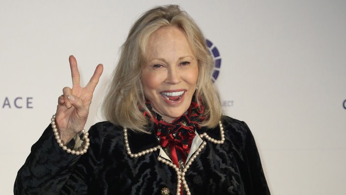 Faye Dunaway lächelt und zeigt ein Peace-Zeichen in die Kamera