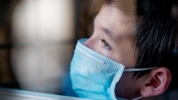 Trauriger Junge mit medizinischer Gesichtsmaske hinter einem Fenster während der Ausganssperre