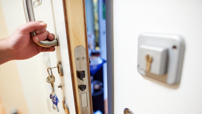 Eine Hand öffnet den Schließmechanismus einer Türe einer forensischen Klinik, der Spalt eröffnet einen unscharfen Blick in das Zimmer.