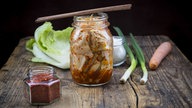 Kimchi: fermentierter, scharfer Kohl. das Bild zeigt diesen in einem Einmachglas auf dem Ess-Stäbchen liegen. Auf dem rustikalen Holztisch liegen außerdem die einzelnen Zutaten, wie Kohl und Chilli.