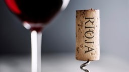 Nahaufnahme eines Kronkorkens auf dem "Rioja" steht, im Hintergrund ein Glas mit Rotwein.