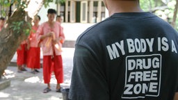 Buddhistisches Drogenentzugskloster Wat Thamkrabok in Thailand: Auf dem Rücken des T-Shirts eines Studenten steht die Aufschrift "my body is a drugfree zone",; im Hintergrund in roten Gewändern sind Mönche des Klosters zu sehen.