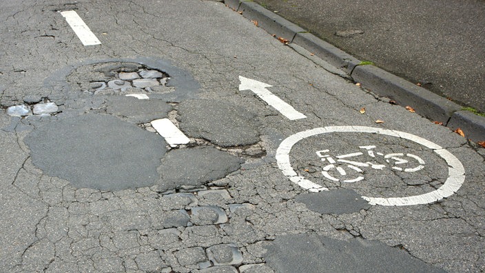 Das Beitragsbild zeigt eine Straße mit vielen Straßenschäden. 