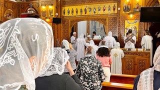 Das Bild zeigt eine koptische Messe im Kloster St.-Antonius. Mitglieder der Gemeinde stehen dabei in einem Kirchenraum mit hölzerner Vertäfelung vor einem Altar.