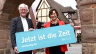 Landesbischof Heinrich Bedford-Strohm und Regionalbischöfin Elisabeth Hann von Weyhern halten das Motto des Kirchentages auf einem Schild: "Jetzt ist die Zeit (Mk 1,15)"