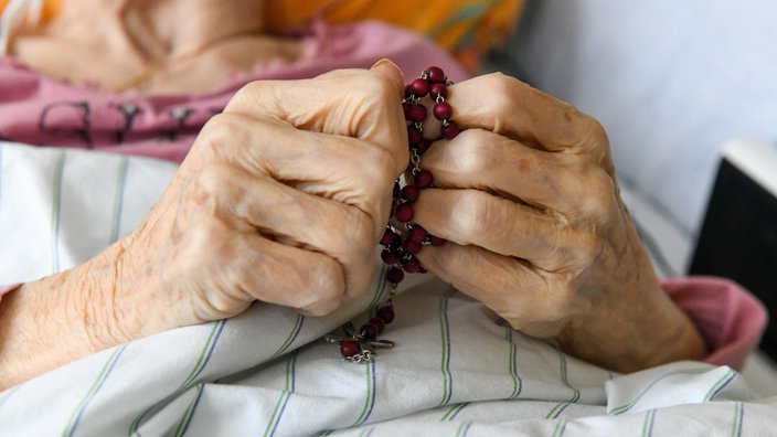 Die Hände einer Frau halten einen Rosenkranz in einem Pflegebett auf der Palliativstation in der Universitätsklinik Bonn am 12. März 2021.