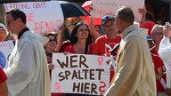  Priester laufen an Teilnehmer:innen der Protestaktion katholischer Frauen im Rahmen von "Maria 2.0"-Aktionen vorbei. Sie halten ein Plakat mit der Aufschrift: "Wer spaltet hier". Die Frauen fordern Geschlechtergerechtigkeit in der katholischen Kirche.