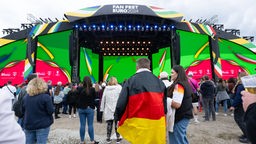 Fans, teilweise mit Deutschlandflagge, stehen vor einer bunten Bühne mit der Aufschrift "Fan Fest Euro 2024"