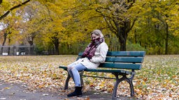 Eine junge Frau sitzt nachdenklich auf einer Bank im Park. 