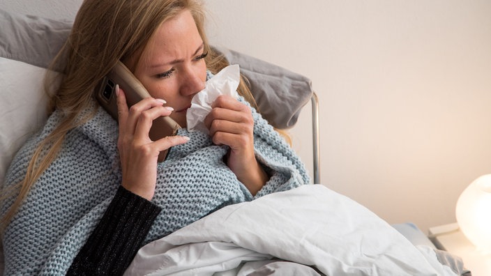 Eine Frau sitzt im Bett mit Taschentusch am Mund und telefoniert