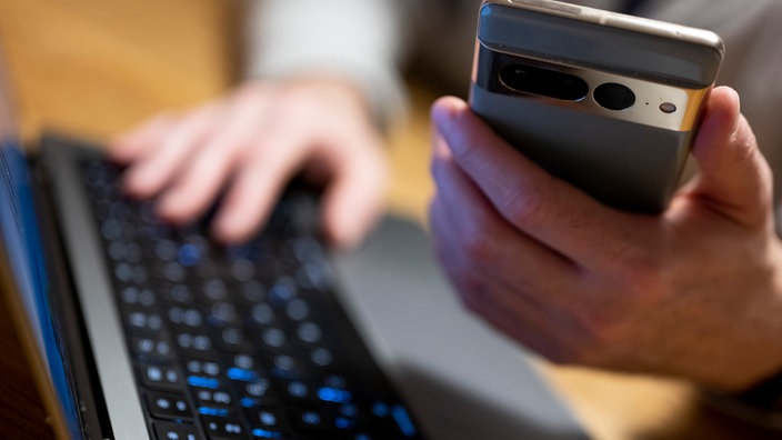 Ein Mann sitzt am Laptop und hält ein Smartphone mit in der Hand. 