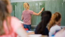 Eine Lehrkraft schreibt in einer Klasse Mathematikaufgaben auf eine Tafel.