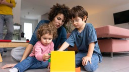 Symbolbild bezahlte Elternschaft: Eine Mutter spielt mit ihrern zwei Kindern; im Hintergrund der Vater.