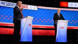 Joe Biden starrt an seinem Rednerpult bei der TV-Debatte um die Präsidentschaftswahl in den USA im us-amerikanischen Fernsehen mit gesenkten Kopf in die Leere, während Trump an seinem Pult in üblicher Manier gestikuliert. 