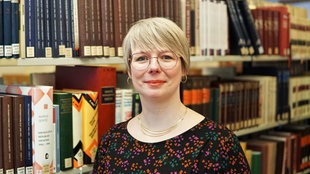 Porträtaufnahme vor einem Bücherregal: Cindy Wittke hat blondes, kürzeres Haar und trägt eine Brille und eine goldene Kette zu ihrem schwarzgemusterten Oberteil.