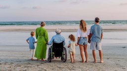 Kinder, Eltern und Großeltern stehen am Strand und blicken aufs Meer. 