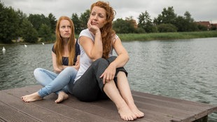 Zwei junge Frauen sitzen auf einem Steg an einem See, ohne sich dabei anzusehen.