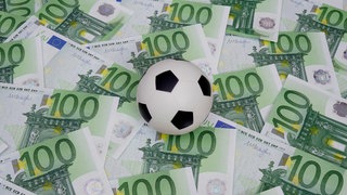 Symbolbild: Geld im Fussball; Auf 100 Euro Geldscheinen liegt ein Fussball.