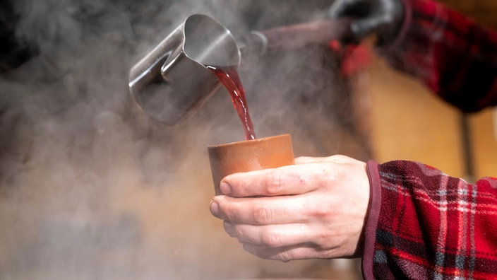 Symbolbild einer Person, die dampfend-heißen Glühwein mit einer Kelle in einen Becher schüttet.