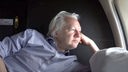 Julian Assange liegt auf der Seite auf seinen Arm gestützt und schaut aus dem Fenster eines Flugzeugs. 