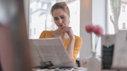 Junge Frau sitzt im Büro am Schreibtisch und liest in der Zeitung. 