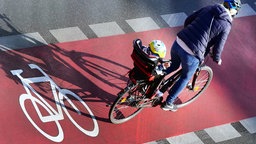 Ein Mutter fährt Fahrrad auf einem roten Radweg, in einem Sitz auf dem Gepäckträger befindet sich ein Kind - beide tragen Helm.