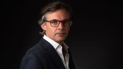 Ein Porträt von Oliver Schlaudt im Anzug, mit nach hinten gekämmtem Haar und Brille vor schwarzem Hintergrund. 