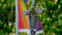 Ein Kreuz steht bei einem Open-Air-Segnungsgottesdienst für Liebende vor der Kirche Christi Auferstehung. Im Hintergrund ist eine Regenbogenfahne zu sehen.