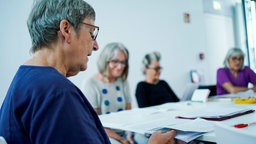 Eine Frau liest weiteren Erwachsenen Menschen in der Stadtbibliothek eine Kurzgeschichte vor.