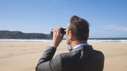 Ein Mann blickt mit einem Fernglas in die Ferne, während er am Strand steht