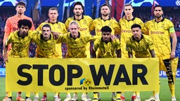 Die Mannschaft von Borussia Dortmund hält ein Banner, darauf steht: Stop War - sponsored by Rheinmetall