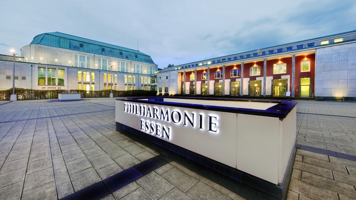 Blick auf das Gebäude der Philharmonie Essen bei Abenddämmerung, mit hell erleuchtetem Schriftzug auf dem Eingangsschild.