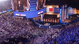 Ein randvoll mit Menschen gefülltes Stadion: auf einem riesigen Bildschirm ist Kamala Harris zu sehen