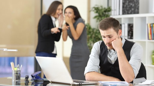 Symbolbild Mobbing am Arbeitsplatz: Kolleginnen reden über am Schreibtisch sitzenden Kollegen.