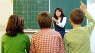 Drei Schüler sitzen einer Lehrerin an der Tafel gegenüber ein Schüler meldet sich