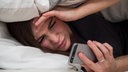 Symbolbild Zeitumstellung: Eine Frau liegt im Bett und schaut müde aufs Smartphone.