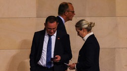 AfD-Spitzenpolitiker Tino Chrupalla und Alice Weidel stehen nebeneinander und blicken auf ihre Handys, im Hintergrund geht CDU-Chef Friedrich Merz vorbei. 