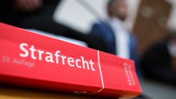 Im Vordergrund ist ein rotes Buch mit dem Titel "Strafrecht" zu sehen, im Hintergrund steht in Unschärfe verschwindend ein Mann, Szene aus dem Gerichtssaal, Symbolbild