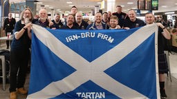 Das Foto zeigt die schottischen Fußballfans am Flughafen. Sie halten eine große Flagge ihres Fanclubs vor sich. 