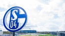 Im Vordergrund dreht sich das Logo des FC Schalke auf der Geschäftsstelle, im Hintergrund steht die Veltins Arena.