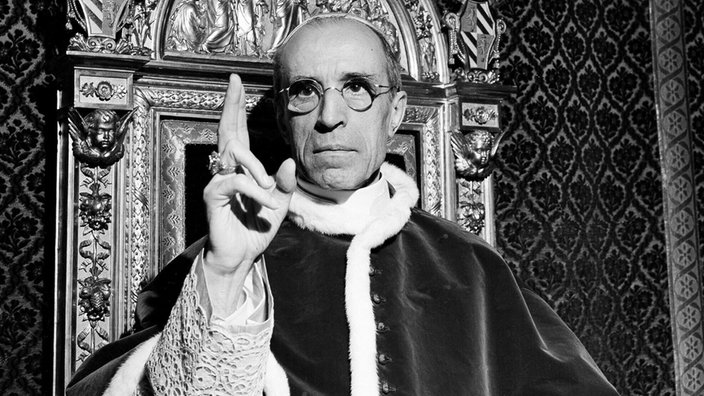 Papst Pius XII. hebt die rechte Hand mit dem Ring des heiligen Petrus, um einen Segen zu sprechen.