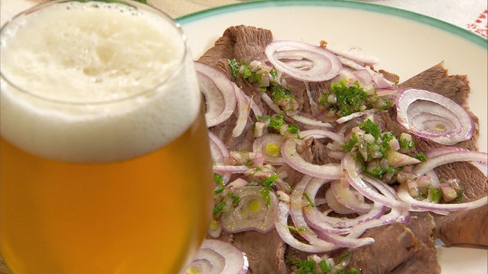 Carpaccio vom Tafelspitz auf einem Teller angerichtet und mit Vinaigrette beträufelt, daneben ein Glas Bier