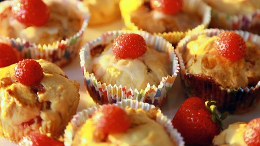 In bunten Muffinförmchen befinden sich kleine, helle Gebäckteilchen, die mit einer Erdbeere garniert sind.