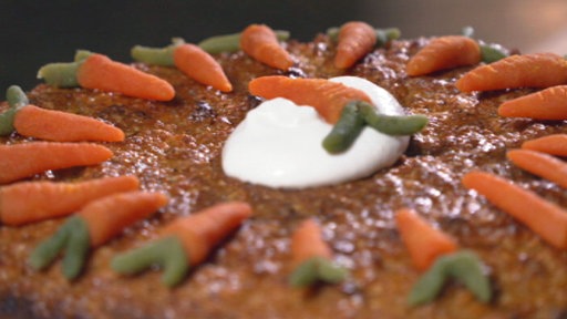 Ein brauner Kuchen verziert mit  kleinen Marzipan-Karotten, in der Mitte ein weißer Klecks, mit einer weiteren kleinen Marzipan-Karotte