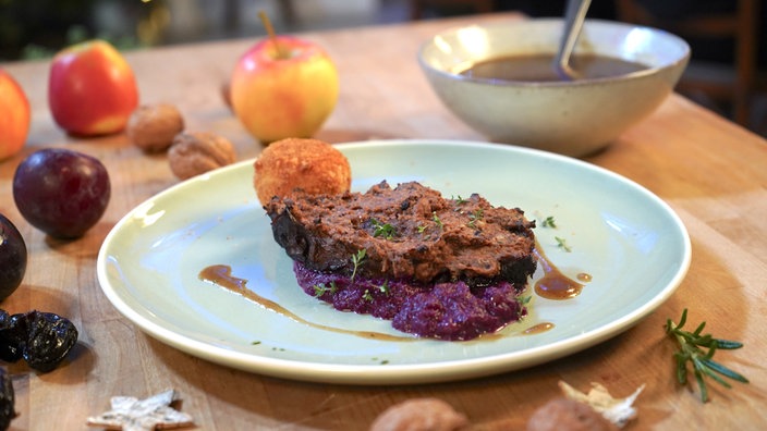 Das Bild zeigt den fertigen Rinderbraten mit Pflaumenkruste. Er wird gemeinsam mit Rotkohl-Püree und Süßkartoffel-Kroketten auf einem Teller angerichtet.